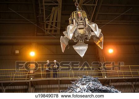 Stock Photo - Steel workers overseeing mechanical grabber in steel foundry. Fotosearch