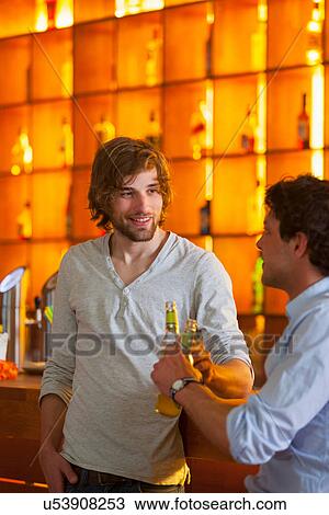 Stock Image - Two men standing at bar with bottles of beer. Fotosearch
