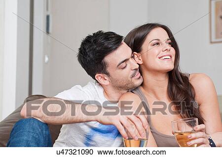 Stock Image - Young couple sitting on sofa sharing glass of wine. Fotosearch