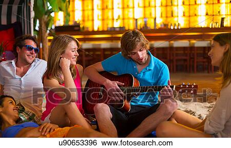 Stock Photograph - Young man playing guitar to friends in bar. Fotosearch