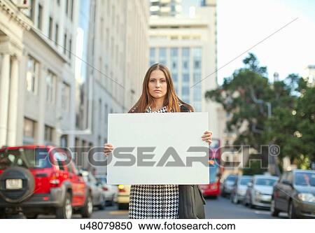 Stock Image - Young woman holding sheet of blank white card on city street. Fotosearch