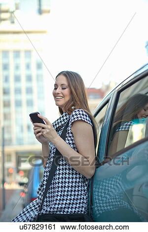Stock Image - Young woman leaning against car using mobile phone. Fotosearch