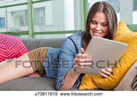 Stock Photo - Young woman lying on sofa holding digital tablet. Fotosearch