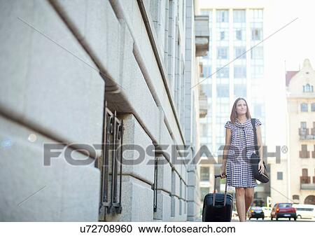 Stock Image - Young woman pulling wheeled case on street. Fotosearch
