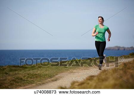 Stock Image - Young woman running along coastal path. Fotosearch