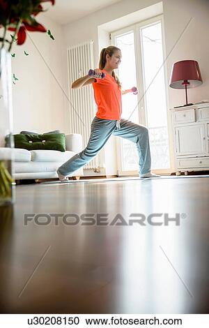 Stock Image - Young woman using dumb-bells at home. Fotosearch