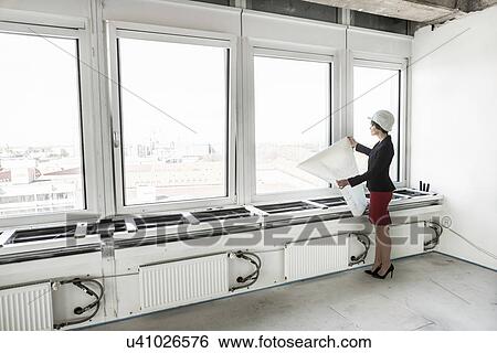 Stock Photograph - Businesswoman standing in empty unfinished office wearing hardhat and looking at plans. Fotosearch