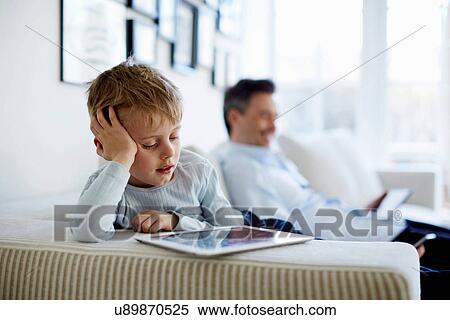 Stock Photography - Father and son sitting on sofa using digital tablets. Fotosearch