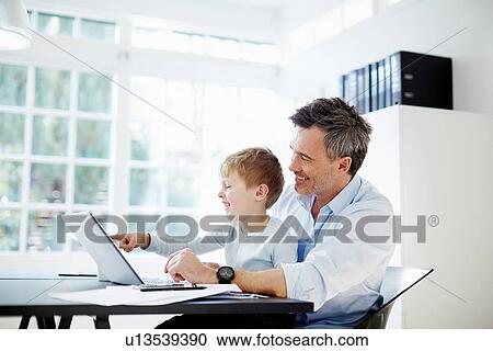 Man sitting at desk with son on his knee with laptop computer and paperwork View Large Photo Image Stock Image - Man sitting at desk with son on his knee with laptop computer and paperwork. Fotosearch