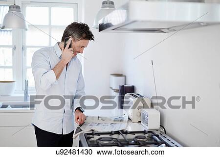 Stock Image - Man standing in kitchen making phone-call and reading newspaper. Fotosearch