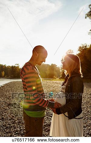 Stock Photograph - Pregnant woman and partner standing face to face beside river holding windmill. Fotosearch