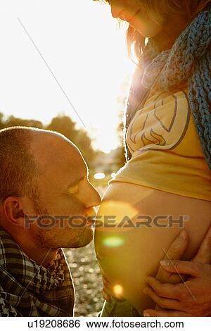 Stock Photograph - Pregnant woman with partner kissing bump. Fotosearch