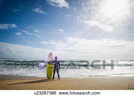 Surfer standing holding surfboard View Large Photo Image Stock Photograph - Surfer standing holding surfboard. Fotosearch