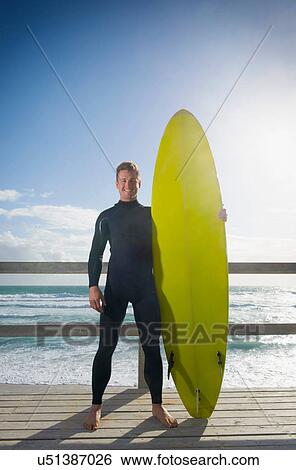 Surfer standing holding surfboard View Large Photo Image Stock Photograph - Surfer standing holding surfboard. Fotosearch
