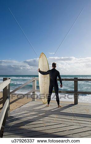 Stock Photo - Surfer with surf board on wooden viewing platform by beach. Fotosearch