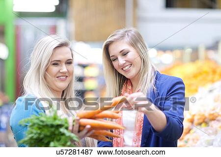 Stock Photo - Two young women checking vegetables in indoor market. Fotosearch
