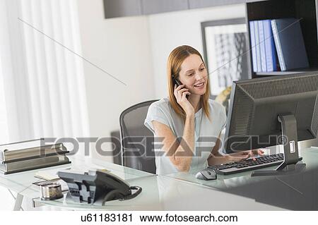 Stock Image - Business woman using computer and phone in office. Fotosearch