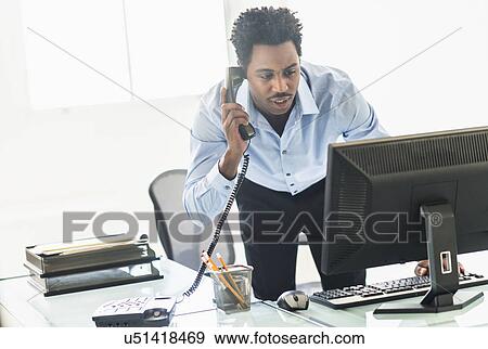 Stock Photo - Businessman working on computer and talking on cell phone. Fotosearch