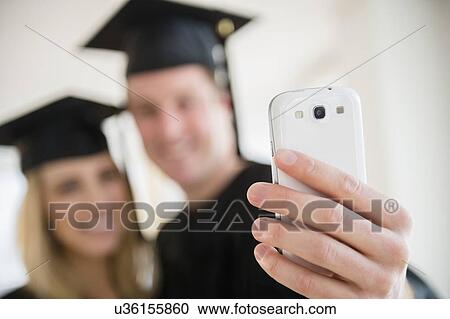 Stock Image - Couple of friends in graduation gowns taking photo with smartphone. Fotosearch