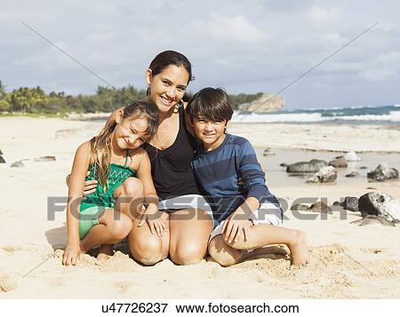 Stock Photo - Portrait of girl (6-7) and boy (10-11) sitting on beach with mother. Fotosearch