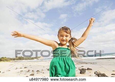 Stock Image - Portrait of girl (6-7) standing on beach with arms raised. Fotosearch