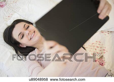 Stock Photo - Portrait of young woman lying down on bed and holding digital tablet. Fotosearch