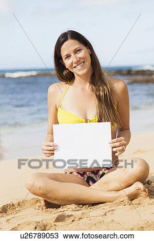 Stock Image - Portrait of young woman with blank paper sitting on beach. Fotosearch