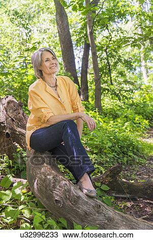 Stock Image - Senior woman sitting on log in forest. Fotosearch