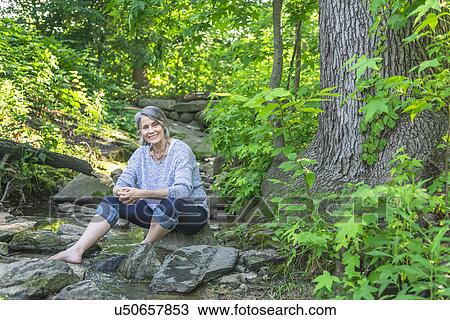 Stock Image - Senior woman sitting on rock in forest. Fotosearch