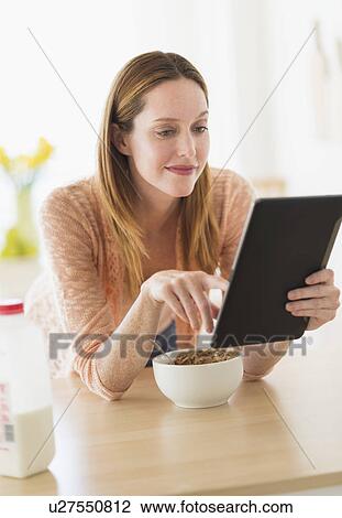 Stock Image - Woman eating breakfast and looking at tablet pc. Fotosearch
