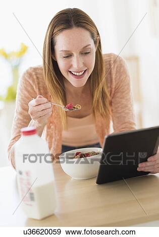 Stock Photo - Woman eating breakfast and looking at tablet pc. Fotosearch
