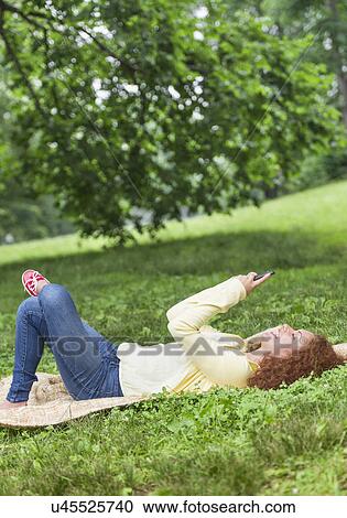 Stock Image - Woman lying on grass in park and using mobile phone. Fotosearch