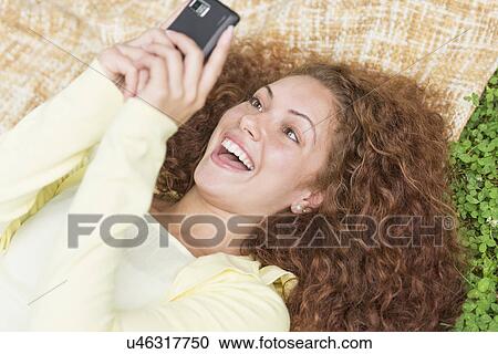 Stock Image - Woman lying on grass in park and using mobile phone. Fotosearch
