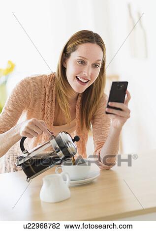 Stock Image - Woman pouring coffee and looking at phone. Fotosearch