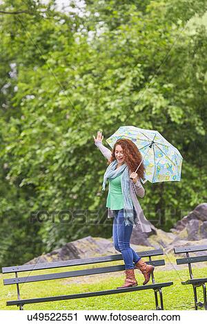 Stock Image - Woman with umbrella dancing on bench in park. Fotosearch