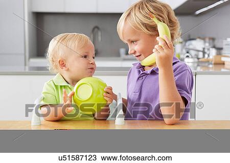 Stock Image - Brothers playing with bowl and banana at table. Fotosearch