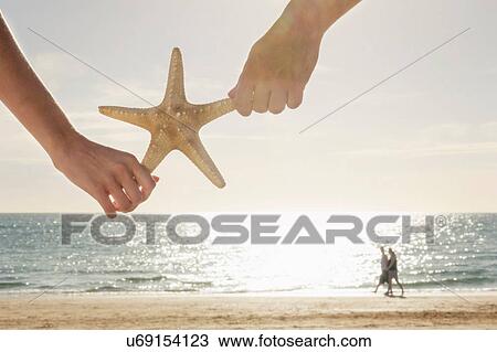 Stock Image - Cropped hands holding starfish, in front of couple walking along beach. Fotosearch