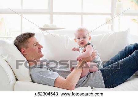 Stock Image - Father lying on sofa holding baby. Fotosearch