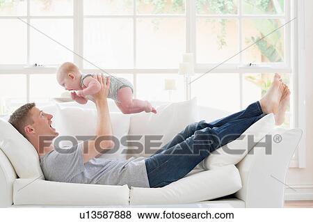 Stock Photo - Father lying on sofa lifting baby. Fotosearch