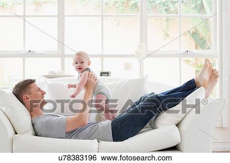 Stock Photograph - Father lying on sofa lifting baby. Fotosearch
