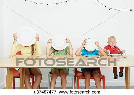 Stock Photo - Four children sitting at table, three covering faces with plates. Fotosearch