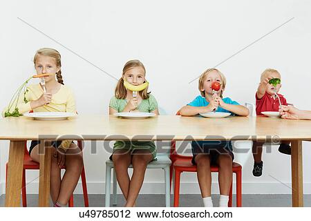 Stock Image - Four children sitting at table with vegetables on forks. Fotosearch