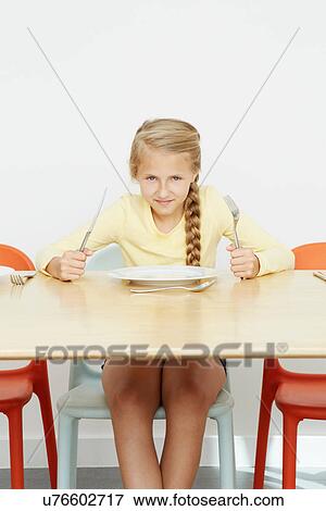 Stock Photo - Girl sitting at table with empty plate holding cutlery. Fotosearch