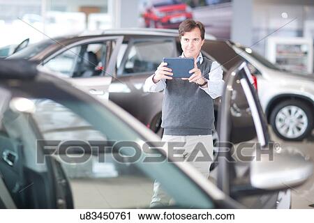 Stock Image - Mid adult man photographing car in showroom. Fotosearch