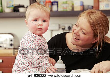 Stock Image - Mother and baby sitting in kitchen with baby bottle. Fotosearch
