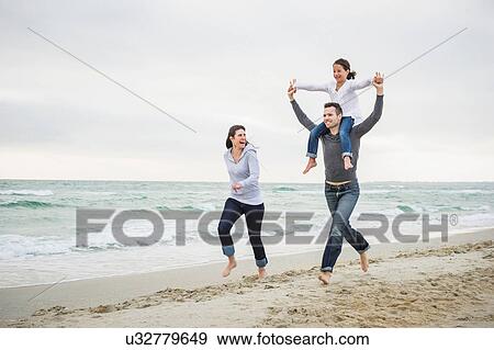 Stock Photo - Mother and father running along beach with girl on shoulders. Fotosearch