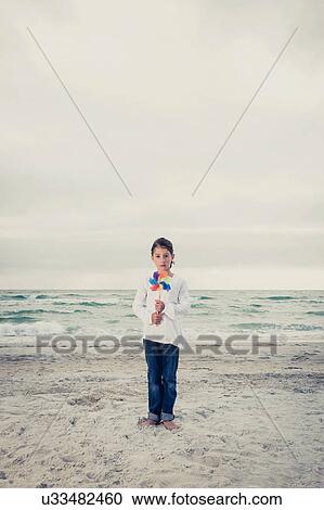 Stock Image - Portrait of girl standing on beach holding windmill. Fotosearch