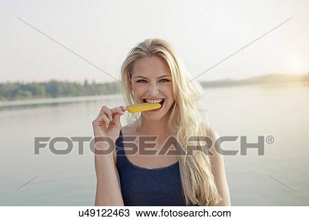 Stock Image - Portrait of young woman biting ice lolly. Fotosearch