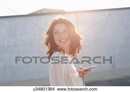 Picture - Portrait of young woman holding cell phone in sunlight. Fotosearch