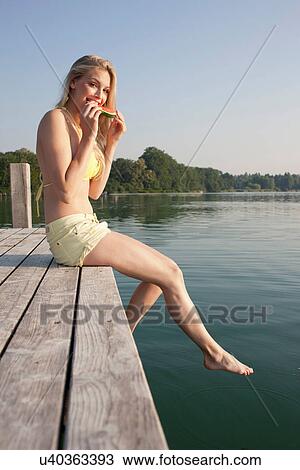 Stock Image - Portrait of young woman on pier eating watermelon. Fotosearch